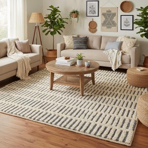 Op de afbeelding: Een woonkamer met een beige bank, een ronde houten salontafel en een patroonkleed. Het kleed heeft een crèmekleurige achtergrond met donkergrijze rechthoekige strepen. Decoratieve items zijn planten, kussens en wandkunst. Een houten vloer en een staande lamp.