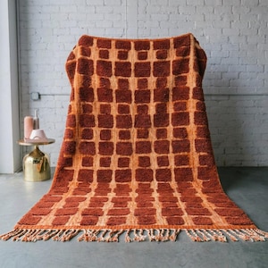Pode incluir: Um cobertor grande e retangular com estampa laranja e marrom com um design de quadrados repetidos. O cobertor tem franjas ao longo da borda inferior e é drapeado sobre uma cadeira. O fundo é uma parede de tijolos brancos.