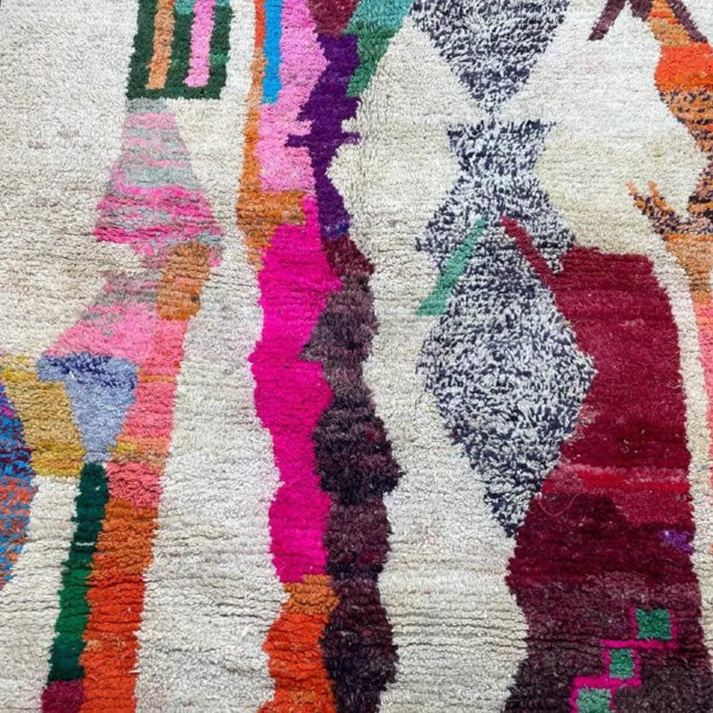 A vibrant, high-pile handmade Moroccan wool rug displayed in a bright industrial studio against a white brick wall. The rug features an eclectic, abstract tribal design with bold splashes of neon pink, teal green, orange, royal blue, and charcoal grey on a natural cream base. The patterns include geometric shapes, abstract figures, and spontaneous color blocking. The rug showcases a thick, plush shaggy texture and authentic hand-knotted cream tassels at the bottom.