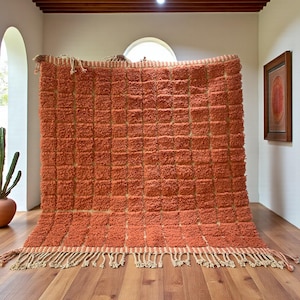 Puede incluir: Una alfombra rectangular naranja con un patrón texturizado en forma de cuadrícula. Tiene un borde crema y flecos en el borde inferior. La alfombra se exhibe en interiores, contra una pared.