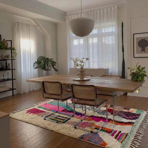 A vibrant, high-pile handmade Moroccan wool rug displayed in a bright industrial studio against a white brick wall. The rug features an eclectic, abstract tribal design with bold splashes of neon pink, teal green, orange, royal blue, and charcoal grey on a natural cream base. The patterns include geometric shapes, abstract figures, and spontaneous color blocking. The rug showcases a thick, plush shaggy texture and authentic hand-knotted cream tassels at the bottom.