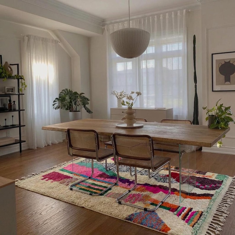 A vibrant, high-pile handmade Moroccan wool rug displayed in a bright industrial studio against a white brick wall. The rug features an eclectic, abstract tribal design with bold splashes of neon pink, teal green, orange, royal blue, and charcoal grey on a natural cream base. The patterns include geometric shapes, abstract figures, and spontaneous color blocking. The rug showcases a thick, plush shaggy texture and authentic hand-knotted cream tassels at the bottom.