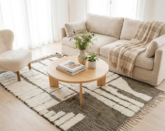 May include: A rectangular area rug featuring a geometric pattern of cream-coloured tufted blocks against a dark grey textured background with fringe on the ends. The rug is placed under a round wooden coffee table, a beige sofa, and a chair.