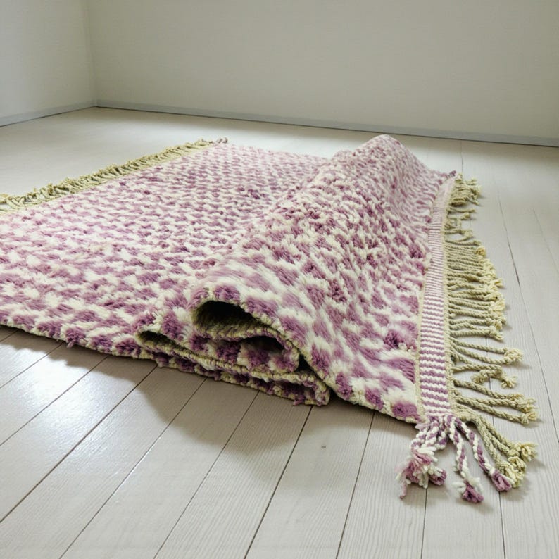 Close-up of a Wistful Mauve Moroccan wool rug handwoven from natural wool by Berber artisans. The rug features a soft mauve and cream color palette with a beautiful geometric pattern and traditional fringes. This thick and plush Moroccan carpet adds warmth, softness, and artisanal charm to living rooms, bedrooms, or boho-chic interiors.