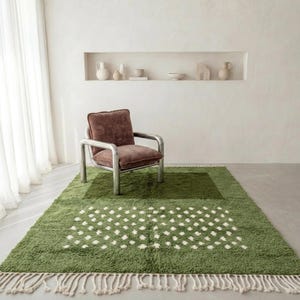 A plush handwoven Moroccan Berber rug in a rich moss green. The thick wool carpet features a solid dark green band near the top and a lighter mossy green base beautifully adorned with small, cream-colored geometric diamond motifs at the bottom. It is draped elegantly against a white painted brick wall next to a gold accent table with trailing ivy, perfectly capturing a warm, earthy, and organic modern interior design aesthetic.