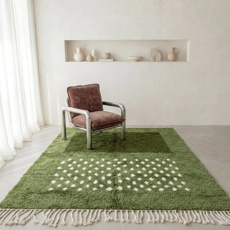 A plush handwoven Moroccan Berber rug in a rich moss green. The thick wool carpet features a solid dark green band near the top and a lighter mossy green base beautifully adorned with small, cream-colored geometric diamond motifs at the bottom. It is draped elegantly against a white painted brick wall next to a gold accent table with trailing ivy, perfectly capturing a warm, earthy, and organic modern interior design aesthetic.