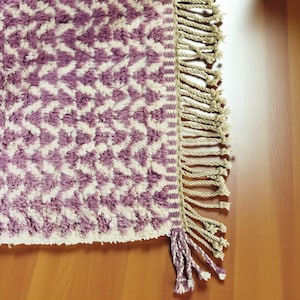 Close-up of a Wistful Mauve Moroccan wool rug handwoven from natural wool by Berber artisans. The rug features a soft mauve and cream color palette with a beautiful geometric pattern and traditional fringes. This thick and plush Moroccan carpet adds warmth, softness, and artisanal charm to living rooms, bedrooms, or boho-chic interiors.