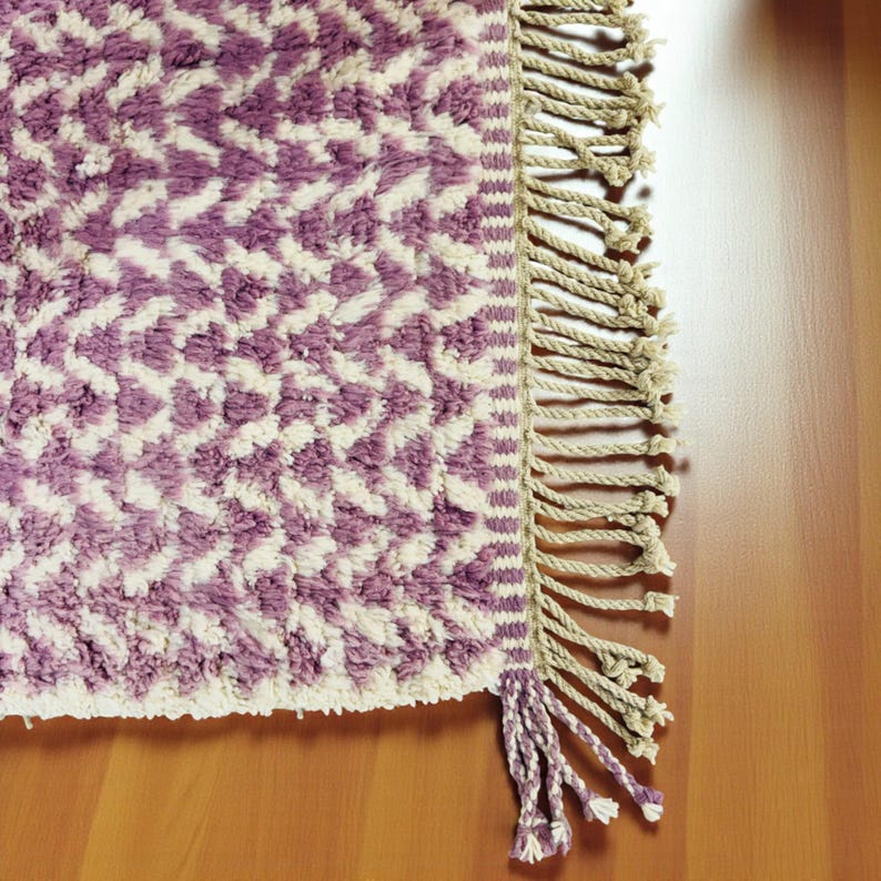 Close-up of a Wistful Mauve Moroccan wool rug handwoven from natural wool by Berber artisans. The rug features a soft mauve and cream color palette with a beautiful geometric pattern and traditional fringes. This thick and plush Moroccan carpet adds warmth, softness, and artisanal charm to living rooms, bedrooms, or boho-chic interiors.