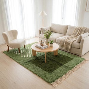 A plush handwoven Moroccan Berber rug in a rich moss green. The thick wool carpet features a solid dark green band near the top and a lighter mossy green base beautifully adorned with small, cream-colored geometric diamond motifs at the bottom. It is draped elegantly against a white painted brick wall next to a gold accent table with trailing ivy, perfectly capturing a warm, earthy, and organic modern interior design aesthetic.
