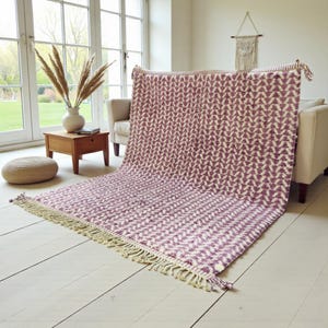 Handwoven Moroccan wool rug in wistful mauve and cream tones. Features a soft geometric pattern with textured pile and hand-knotted fringes. Perfect for living rooms, bedrooms, or boho interiors. Made from 100% natural wool by Berber artisans in Morocco. This handmade rug adds warmth, elegance, and Moroccan authenticity to modern or vintage-style spaces.