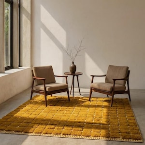 Large handmade Moroccan high-pile shag rug in saturated Thai curry mustard-gold, displayed vertically against a white brick wall. Surface features a tone-on-tone square grid pattern formed through pile variation — raised mustard squares separated by recessed ivory cream channel lines across the entire field. Dense, plush, curly wool pile texture throughout. Knotted braided ivory fringe along the top hem edge and long hand-tied ivory fringe along the bottom edge.