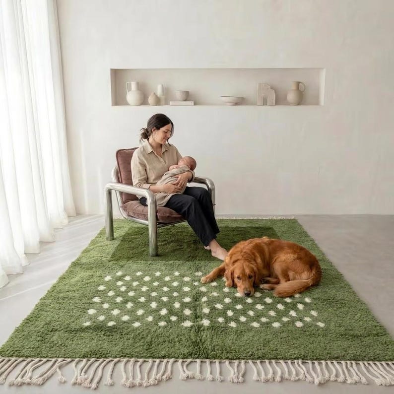 Handmade Moroccan Boujaad rug in soft sage green with scattered ivory white dot motifs across the lower field. Thick plush shaggy wool pile with natural white knotted fringe. Styled in a serene minimalist room with a woman holding a newborn baby in a linen chair, a golden retriever lying on the rug, white curtains and built-in ceramic shelf display. Hand-knotted by Berber artisans in Marrakech, Morocco. A gentle, calming statement rug for nurseries, family rooms and boho interiors.