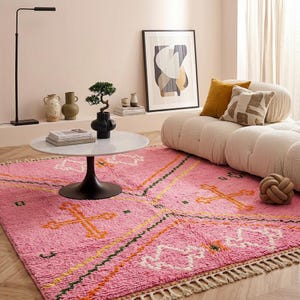 A plush, handwoven Moroccan Boujad rug featuring a vibrant hot pink base adorned with neon orange crosses, white diamonds, and green zigzag geometric patterns. The brightly colored wool carpet is draped elegantly against a white painted brick wall next to a gold accent table with pink pillar candles, perfectly capturing a playful maximalist and dopamine decor aesthetic.