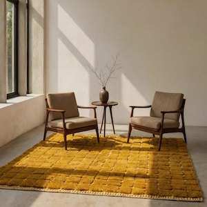 Large handmade Moroccan high-pile shag rug in saturated Thai curry mustard-gold, displayed vertically against a white brick wall. Surface features a tone-on-tone square grid pattern formed through pile variation — raised mustard squares separated by recessed ivory cream channel lines across the entire field. Dense, plush, curly wool pile texture throughout. Knotted braided ivory fringe along the top hem edge and long hand-tied ivory fringe along the bottom edge.