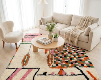May include: A rectangular area rug featuring a cream-coloured base with a multi-coloured geometric pattern. The design includes black lines, pink stripes, orange leaf-like shapes, and a checkered section in green, orange, and black. It has fringe edges.