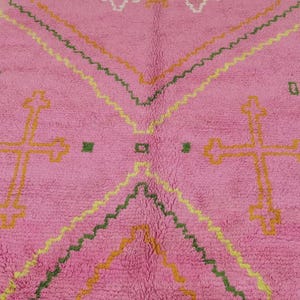 A plush, handwoven Moroccan Boujad rug featuring a vibrant hot pink base adorned with neon orange crosses, white diamonds, and green zigzag geometric patterns. The brightly colored wool carpet is draped elegantly against a white painted brick wall next to a gold accent table with pink pillar candles, perfectly capturing a playful maximalist and dopamine decor aesthetic.