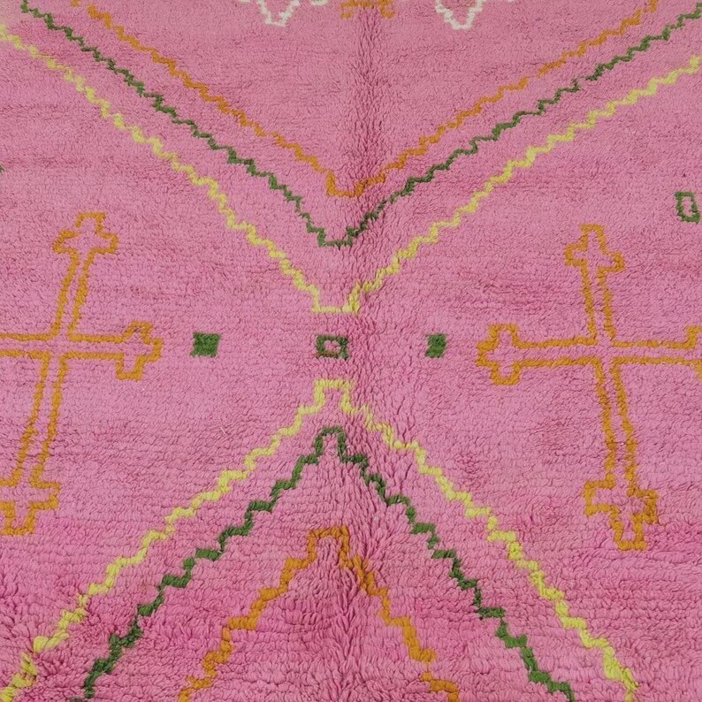 A plush, handwoven Moroccan Boujad rug featuring a vibrant hot pink base adorned with neon orange crosses, white diamonds, and green zigzag geometric patterns. The brightly colored wool carpet is draped elegantly against a white painted brick wall next to a gold accent table with pink pillar candles, perfectly capturing a playful maximalist and dopamine decor aesthetic.