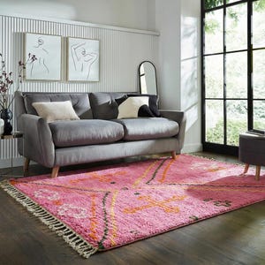 A plush, handwoven Moroccan Boujad rug featuring a vibrant hot pink base adorned with neon orange crosses, white diamonds, and green zigzag geometric patterns. The brightly colored wool carpet is draped elegantly against a white painted brick wall next to a gold accent table with pink pillar candles, perfectly capturing a playful maximalist and dopamine decor aesthetic.