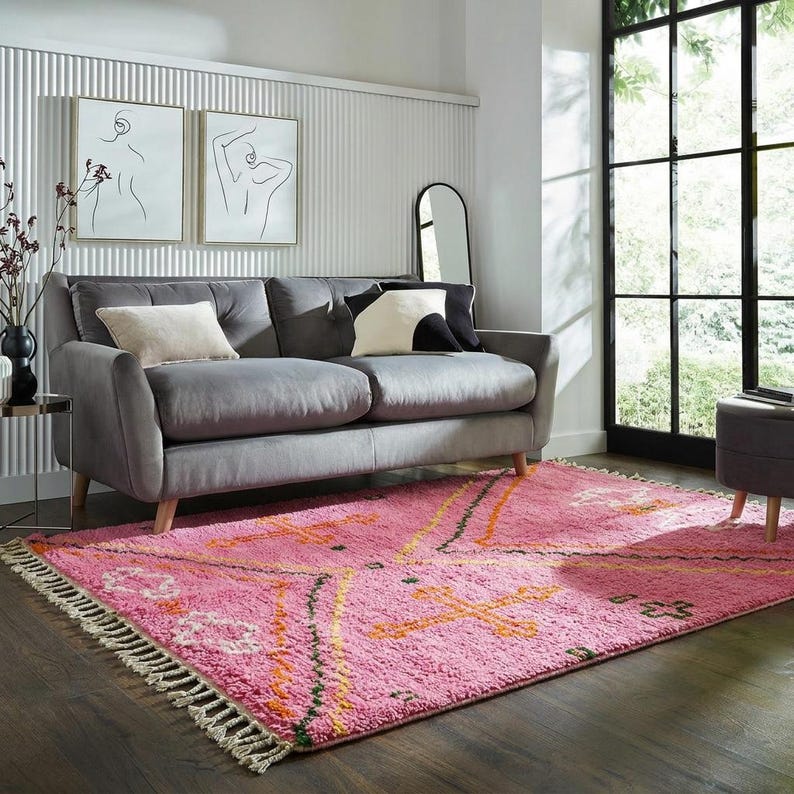 A plush, handwoven Moroccan Boujad rug featuring a vibrant hot pink base adorned with neon orange crosses, white diamonds, and green zigzag geometric patterns. The brightly colored wool carpet is draped elegantly against a white painted brick wall next to a gold accent table with pink pillar candles, perfectly capturing a playful maximalist and dopamine decor aesthetic.