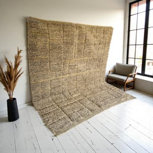 Könnte beinhalten: Ein großer, rechteckiger Teppich mit einem strukturierten, geometrischen Muster in Beige- und Brauntönen. Der Teppich wird vor einer weißen Wand präsentiert, mit einer schwarzen Vase mit getrockneten Pflanzen und einem Holzstuhl im Hintergrund. Das Design des Teppichs weist ein gitterartiges Muster auf.