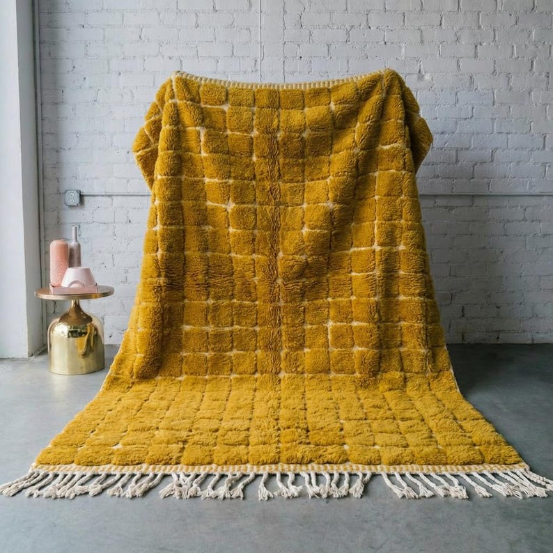 Large handmade Moroccan high-pile shag rug in saturated Thai curry mustard-gold, displayed vertically against a white brick wall. Surface features a tone-on-tone square grid pattern formed through pile variation — raised mustard squares separated by recessed ivory cream channel lines across the entire field. Dense, plush, curly wool pile texture throughout. Knotted braided ivory fringe along the top hem edge and long hand-tied ivory fringe along the bottom edge.