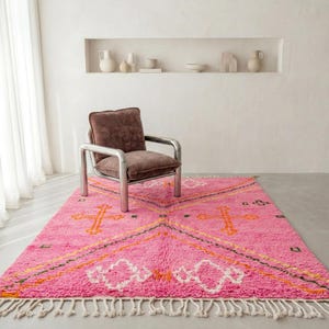 A plush, handwoven Moroccan Boujad rug featuring a vibrant hot pink base adorned with neon orange crosses, white diamonds, and green zigzag geometric patterns. The brightly colored wool carpet is draped elegantly against a white painted brick wall next to a gold accent table with pink pillar candles, perfectly capturing a playful maximalist and dopamine decor aesthetic.