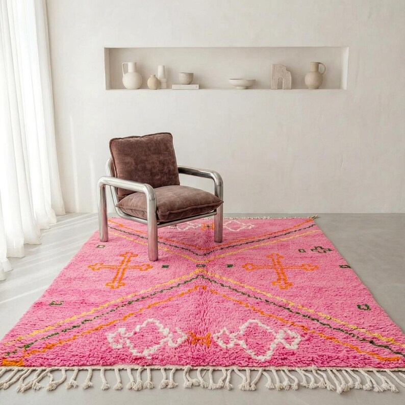 A plush, handwoven Moroccan Boujad rug featuring a vibrant hot pink base adorned with neon orange crosses, white diamonds, and green zigzag geometric patterns. The brightly colored wool carpet is draped elegantly against a white painted brick wall next to a gold accent table with pink pillar candles, perfectly capturing a playful maximalist and dopamine decor aesthetic.