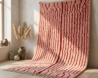 Può includere: Un tappeto rettangolare a pelo lungo con un motivo di linee verticali ondulate rosso scuro su sfondo rosa tenue. Il tappeto presenta un bordo con frange sul lato inferiore ed è esposto in una posizione verticale e drappeggiata.