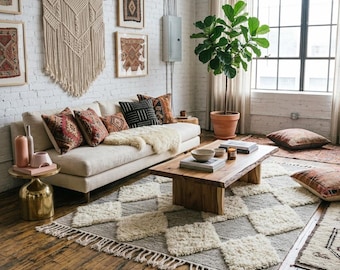 May include: A cream-coloured sofa with patterned throw pillows and a white faux fur throw, a wooden coffee table, a grey and white diamond-patterned rug with fringe, a gold side table, and a large potted fiddle leaf fig plant in a room.