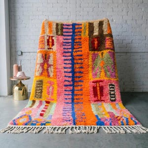 May include: A colourful rug with a geometric design. The rug features orange, pink, blue, and brown shades. It has a shaggy texture and a fringe along the bottom edge. The rug is displayed against a white brick wall.