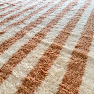 Elegant handwoven Moroccan rug with vertical stripes in Cameo Rose and soft white. Perfect for boho, coastal, minimalist, or Japandi-inspired spaces. Made from natural wool, this artisan piece blends texture and comfort for your living room, bedroom, or entryway. A modern pastel rug with warm earthy tones and timeless Amazigh character. Custom sizes available.