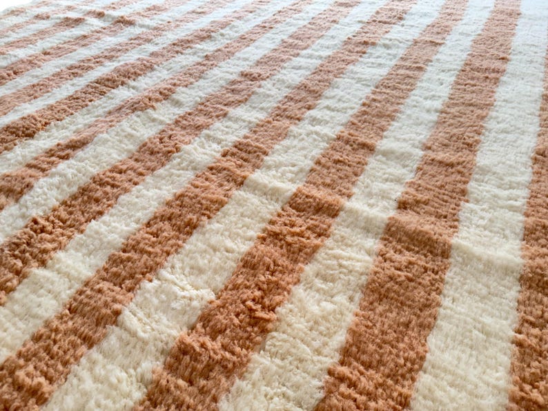 Elegant handwoven Moroccan rug with vertical stripes in Cameo Rose and soft white. Perfect for boho, coastal, minimalist, or Japandi-inspired spaces. Made from natural wool, this artisan piece blends texture and comfort for your living room, bedroom, or entryway. A modern pastel rug with warm earthy tones and timeless Amazigh character. Custom sizes available.