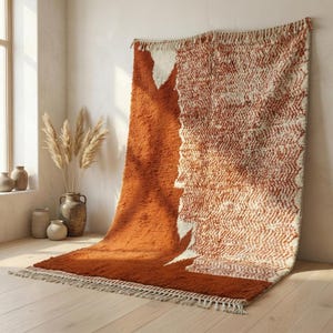 Op de afbeelding: Een groot, rechthoekig vloerkleed met een textuur, geometrisch ontwerp. Het vloerkleed heeft een combinatie van roestoranje en crèmekleuren. Het vloerkleed is gemaakt van een zacht materiaal en heeft een franje langs de onderrand. Het vloerkleed wordt op een lichte houten vloer getoond.