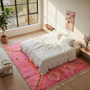 A plush, handwoven Moroccan Boujad rug featuring a vibrant hot pink base adorned with neon orange crosses, white diamonds, and green zigzag geometric patterns. The brightly colored wool carpet is draped elegantly against a white painted brick wall next to a gold accent table with pink pillar candles, perfectly capturing a playful maximalist and dopamine decor aesthetic.