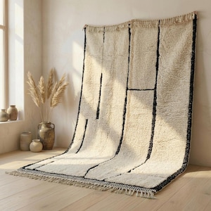 May include: A cream-colored rug with a geometric black design. The rug has a thick, textured pile and a fringe along the bottom edge. The rug is displayed against a neutral wall and wooden floor, creating a cozy and inviting atmosphere.