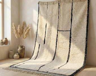Può includere: Un tappeto color crema con un design geometrico nero. Il tappeto ha un pelo spesso e strutturato e un bordo con frange. È esposto contro un muro neutro e un pavimento in legno, creando un'atmosfera calda e accogliente.