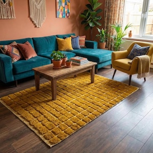 Handmade Moroccan Boujaad rug in rich mustard yellow featuring an all-over raised check square pattern with pronounced high-low carved pile texture. Natural ivory fringe. Styled in a bold maximalist boho living room with a teal velvet sectional sofa, mustard yellow armchair, terracotta painted walls, macrame wall hanging, tropical plants and rustic wood coffee table with terracotta pots. Hand-knotted by Berber artisans in Marrakech, Morocco. A warm statement rug for colorful boho interiors.