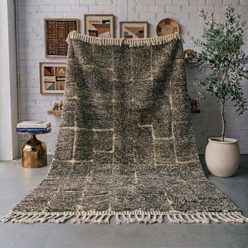 Handmade Moroccan wool rug in warm Travertine and beige tones, featuring an abstract irregular grid and block pattern with a richly textured high pile. Natural lighter lines separate the earthy beige and taupe sections, creating a modern architectural look. Finished with handwoven striped edges and natural fringes, the rug is styled in a calm neutral room with ceramic pottery and greenery. A contemporary Berber rug ideal for organic modern, Japandi, minimalist, and wabi-sabi interiors.