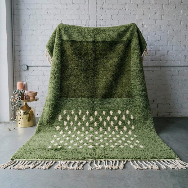A plush handwoven Moroccan Berber rug in a rich moss green. The thick wool carpet features a solid dark green band near the top and a lighter mossy green base beautifully adorned with small, cream-colored geometric diamond motifs at the bottom. It is draped elegantly against a white painted brick wall next to a gold accent table with trailing ivy, perfectly capturing a warm, earthy, and organic modern interior design aesthetic.
