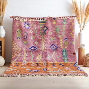 May include: A rectangular, hand-woven rug with a mauve background and geometric patterns in yellow, blue, and green. The rug features an orange border with fringe detailing along the bottom edge. The rug is displayed against a white wall.