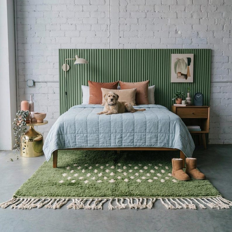 A plush handwoven Moroccan Berber rug in a rich moss green. The thick wool carpet features a solid dark green band near the top and a lighter mossy green base beautifully adorned with small, cream-colored geometric diamond motifs at the bottom. It is draped elegantly against a white painted brick wall next to a gold accent table with trailing ivy, perfectly capturing a warm, earthy, and organic modern interior design aesthetic.