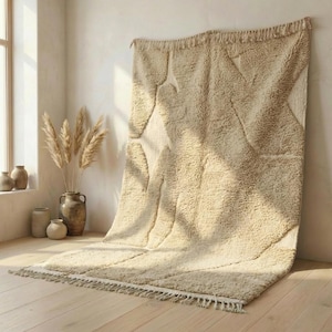Op de afbeelding: Een groot, beige tapijt met een geometrisch ontwerp is tegen een muur gedrapeerd. Het tapijt heeft een dikke, getextureerde pool en een franje langs de onderrand. Het ontwerp heeft grote, abstracte vormen in een lichtere tint beige. Het tapijt wordt in een goed verlichte kamer getoond.