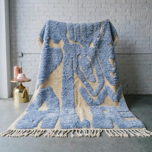 Peut inclure: Un grand tapis texturé avec un motif abstrait bleu clair et beige. Le tapis a une texture touffue et une frange sur le bord inférieur. Le tapis est drapé sur une chaise, mettant en valeur son design et sa texture.