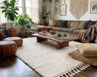 May include: A living room featuring a large cream-coloured shag rug with fringe, a low wooden coffee table, a brown velvet sectional sofa with patterned throw pillows, two leather poufs, and several potted green plants near a window.