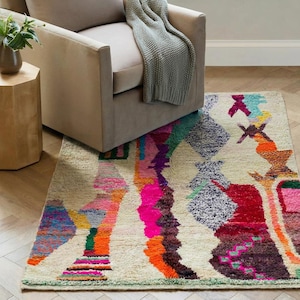 A vibrant, high-pile handmade Moroccan wool rug displayed in a bright industrial studio against a white brick wall. The rug features an eclectic, abstract tribal design with bold splashes of neon pink, teal green, orange, royal blue, and charcoal grey on a natural cream base. The patterns include geometric shapes, abstract figures, and spontaneous color blocking. The rug showcases a thick, plush shaggy texture and authentic hand-knotted cream tassels at the bottom.