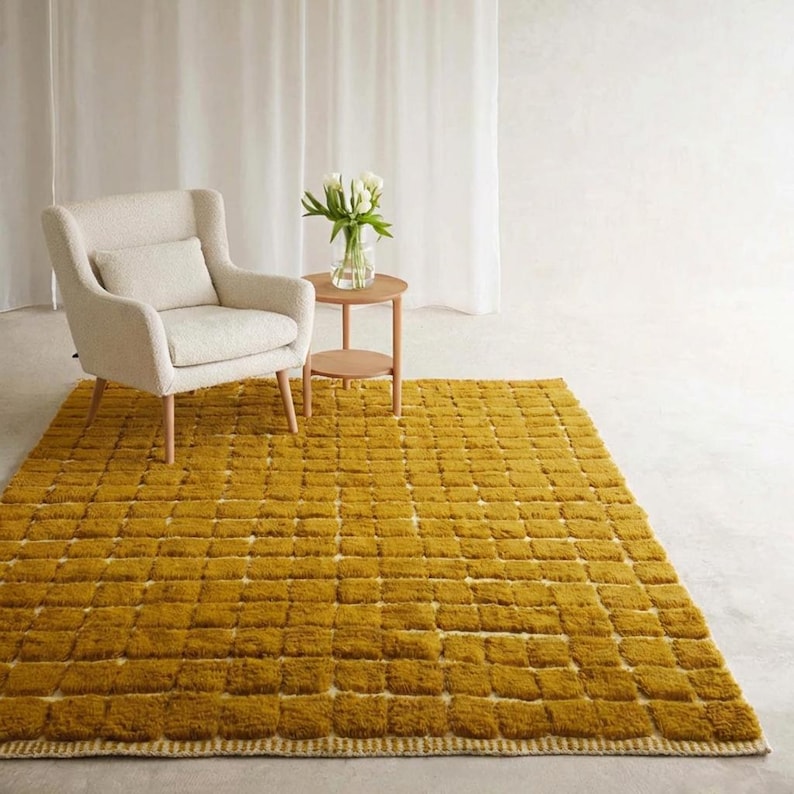 Large handmade Moroccan high-pile shag rug in saturated Thai curry mustard-gold, displayed vertically against a white brick wall. Surface features a tone-on-tone square grid pattern formed through pile variation — raised mustard squares separated by recessed ivory cream channel lines across the entire field. Dense, plush, curly wool pile texture throughout. Knotted braided ivory fringe along the top hem edge and long hand-tied ivory fringe along the bottom edge.