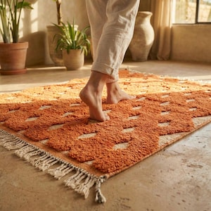 May include: A rectangular rug in shades of orange and cream, featuring a geometric design and fringed edges. The rug has a textured, tactile surface. A person is standing on the rug.