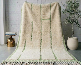 May include: A rectangular area rug featuring a thick, cream-coloured pile with vertical and horizontal green line patterns. The rug has a fringed edge along the bottom and is displayed draped over a surface, showcasing its textured, soft appearance.