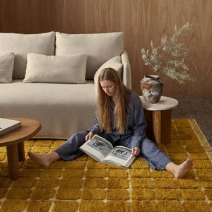 Large handmade Moroccan high-pile shag rug in saturated Thai curry mustard-gold, displayed vertically against a white brick wall. Surface features a tone-on-tone square grid pattern formed through pile variation — raised mustard squares separated by recessed ivory cream channel lines across the entire field. Dense, plush, curly wool pile texture throughout. Knotted braided ivory fringe along the top hem edge and long hand-tied ivory fringe along the bottom edge.