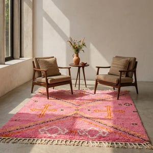 A plush, handwoven Moroccan Boujad rug featuring a vibrant hot pink base adorned with neon orange crosses, white diamonds, and green zigzag geometric patterns. The brightly colored wool carpet is draped elegantly against a white painted brick wall next to a gold accent table with pink pillar candles, perfectly capturing a playful maximalist and dopamine decor aesthetic.